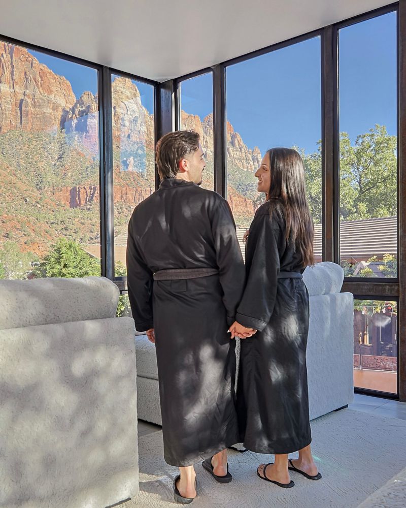 Two guests in robes holding hands at a corner window with red rock canyon views